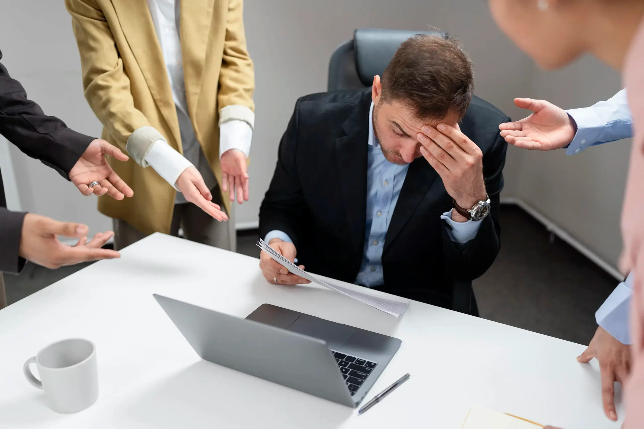 Un employé, submergé par le stress, se tient la tête devant son ordinateur portable tandis que ses collègues semblent lui faire des reproches. Cette scène illustre les risques psychosociaux en entreprise, mettant en lumière l’importance de la prévention et de la gestion du bien-être au travail pour éviter le burnout et les tensions professionnelles.